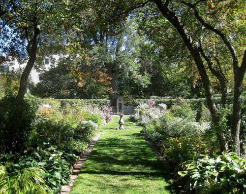 A lush garden pathway lined with flowers and trees, leading to a statue in a serene, green setting.