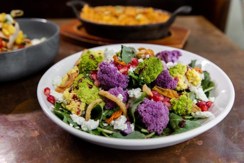 A vibrant salad featuring colorful cauliflower, greens, pomegranate seeds, and crispy toppings in a white bowl.