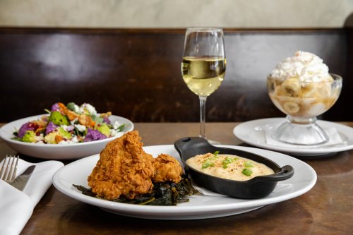 A plate with fried chicken, creamy grits, a salad, a glass of white wine, and a dessert with bananas and whipped cream.