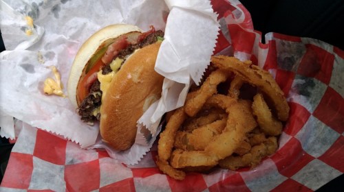 A cheeseburger with lettuce, tomato, and pickles next to a serving of crispy onion rings in a red-checkered paper.