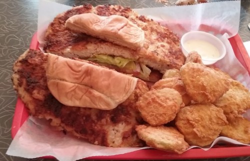 A large breaded sandwich with lettuce and tomato, served with a side of golden fried bites and dipping sauce.