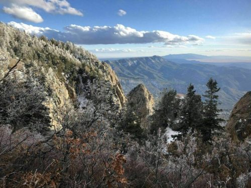 A scenic mountain view with snow-covered trees and rocky cliffs under a blue sky with fluffy clouds.