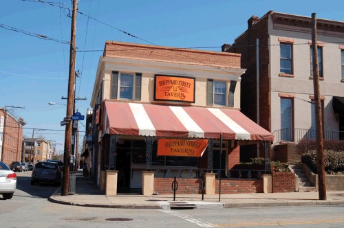 A brick building with a striped awning, featuring a sign that reads "Sheppard Street Tavern" on a sunny day.