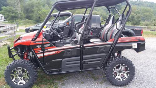 A red and black Kawasaki Teryx 4x4 off-road vehicle parked on gravel, with a scenic background of greenery.