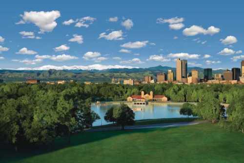 A scenic view of a city skyline with mountains in the background, featuring a lake and lush green parkland.