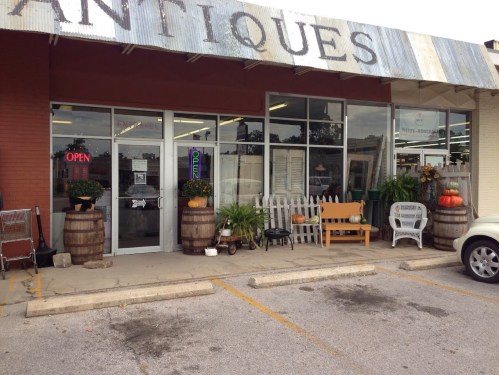 Exterior of an antique shop with barrels, plants, and a sign that says "OPEN" in the window.