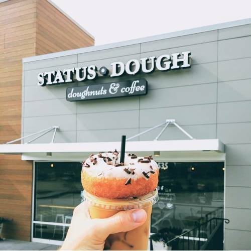 A hand holds a donut topped with chocolate sprinkles in front of the Status Dough shop, featuring a coffee drink.