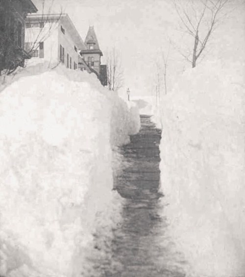 A narrow path cleared through deep snow, flanked by high snowbanks, with buildings visible in the background.