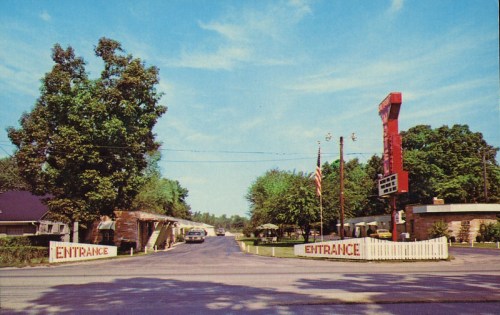 A sunny view of a motel entrance with a sign, trees, and a vintage car parked on a quiet street.