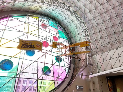A vintage airplane model hangs in a colorful, geometric glass atrium with a "Take Flight" banner.