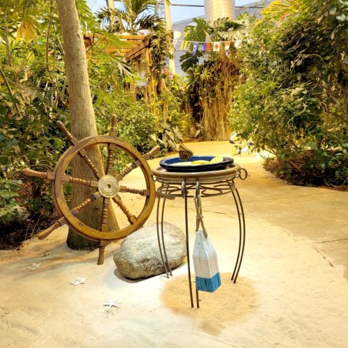 A wooden ship's wheel beside a decorative table with a blue bowl, surrounded by lush greenery and sandy pathways.