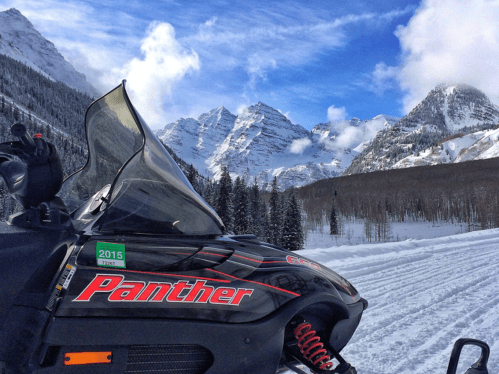 A snowmobile parked on a snowy trail with majestic mountains and a blue sky in the background.