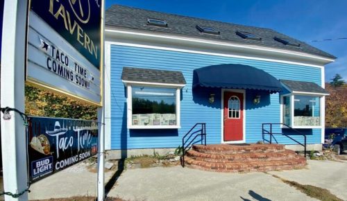 A blue building with a red door and awning, featuring signs for a tavern and an upcoming taco restaurant.