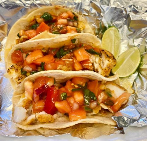 Three fish tacos topped with diced tomatoes and cilantro, served with lime wedges on a foil-lined plate.
