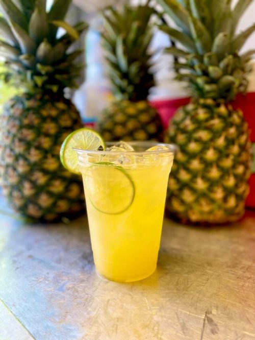 A refreshing yellow drink with ice and a lime slice, in front of three whole pineapples.