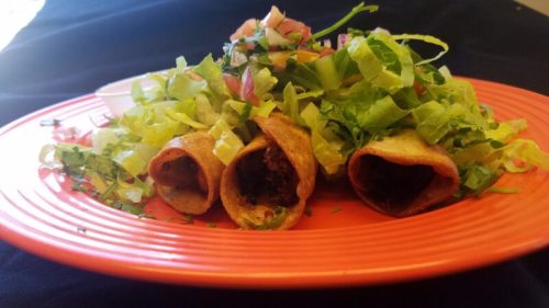 Three crispy taquitos filled with meat, topped with fresh lettuce, diced tomatoes, and a side of sauce on an orange plate.