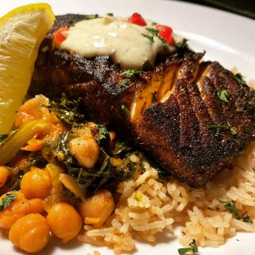 A plate of seasoned fish with a lemon wedge, served over rice and accompanied by chickpeas and greens.