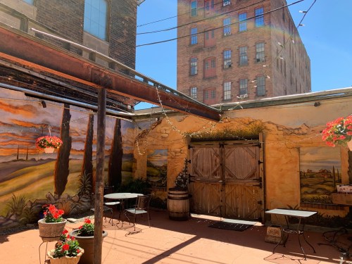 A sunny outdoor patio with a mural, potted flowers, and tables, surrounded by brick buildings and string lights.