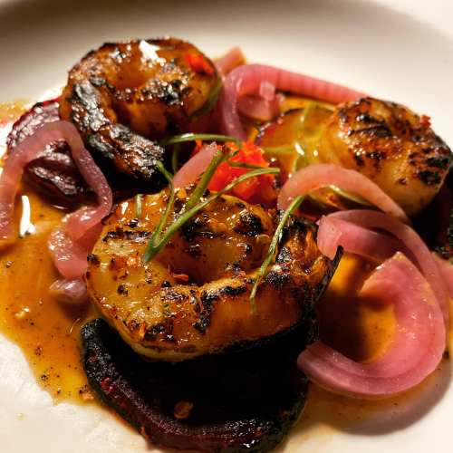 Grilled shrimp with vibrant sauce, garnished with pickled onions and herbs on a white plate.