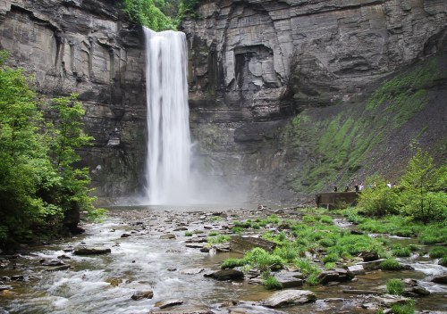 A majestic waterfall cascades down rocky cliffs, surrounded by lush greenery and a serene river below.