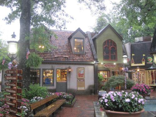 Charming storefronts in a quaint village, surrounded by lush greenery and colorful flowers, on a rainy day.