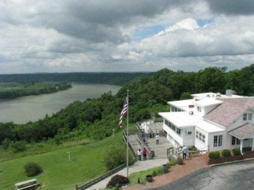 A scenic view of a river valley from a hilltop, featuring a building and an American flag in the foreground.