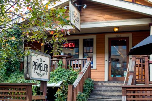 A cozy restaurant entrance with wooden steps, signs reading "The Rambler" and "Open," surrounded by greenery.