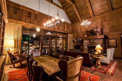 Cozy rustic living room with wooden beams, a stone fireplace, and a large dining table surrounded by leather chairs.