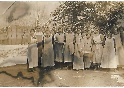 A historical black-and-white photo of a group of women in aprons standing outdoors, with a building and trees in the background.