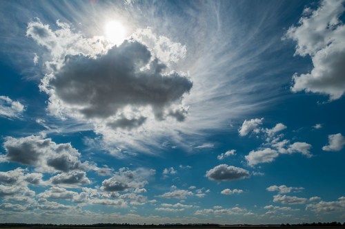 A bright sun peeks through fluffy clouds in a vibrant blue sky, with wispy clouds scattered throughout.