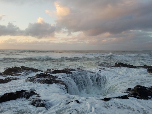 Waves crash against rocky shore at sunset, creating a dramatic scene with foamy water and cloudy sky.