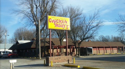 Sign for Chicken Mary's restaurant with a brick building and trees in the background on a clear day.