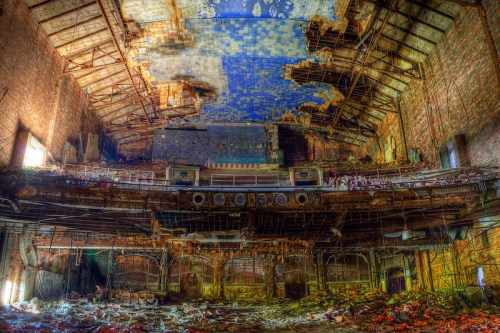 Abandoned theater interior with crumbling walls, a damaged ceiling, and scattered debris on the floor.