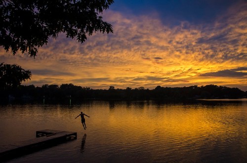 A person jumps off a dock into a calm lake at sunset, with vibrant clouds reflecting on the water.