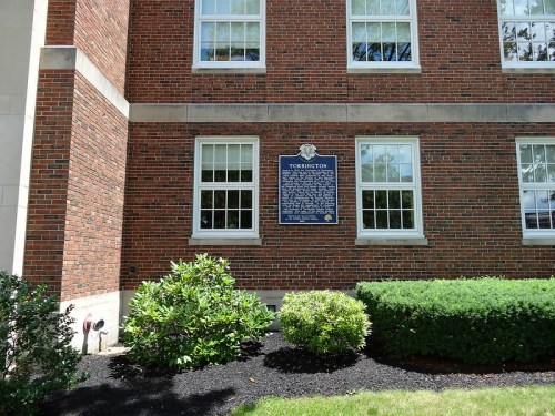 Historical plaque on a brick building, surrounded by green shrubs and trees, with windows visible above.