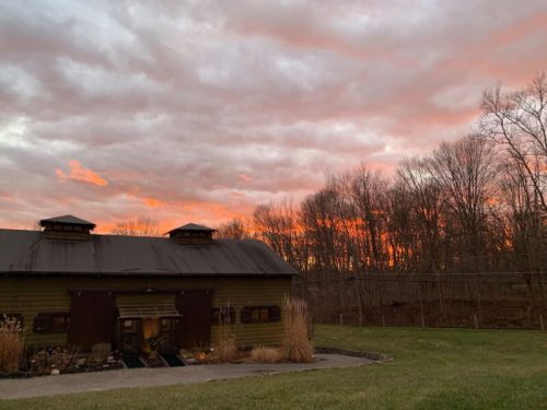 A rustic building with a dark roof against a vibrant sunset sky, surrounded by trees and grassy land.