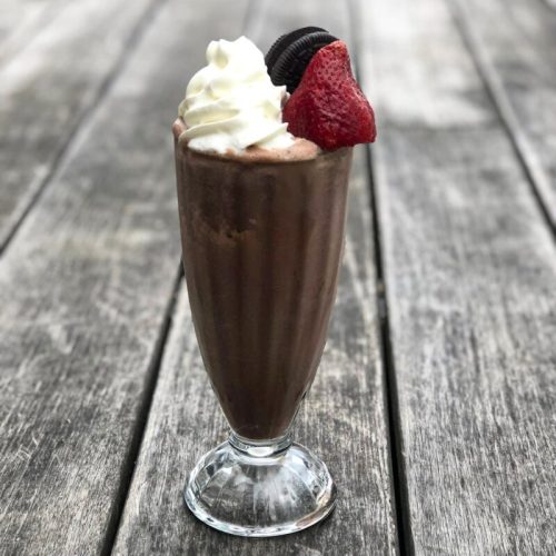 A chocolate milkshake topped with whipped cream, a strawberry, and an Oreo cookie, served in a tall glass on a wooden table.