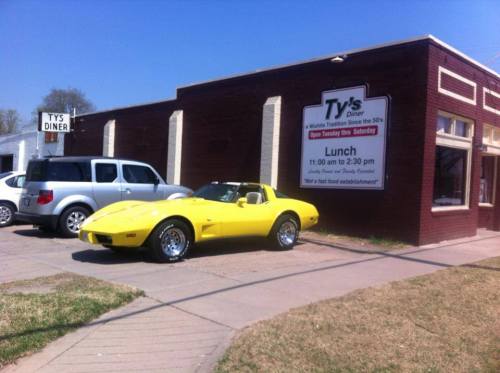 A bright yellow vintage car parked outside Ty's Diner, a retro-style restaurant with a sign displaying lunch hours.