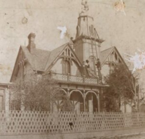Historic house with a tower, featuring a ladder and workers on the roof, surrounded by a picket fence and trees.