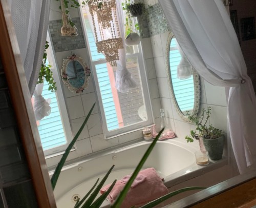A cozy bathroom with a bathtub, plants, and decorative mirrors, featuring soft curtains and natural light.