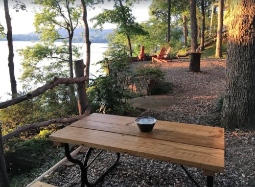 A wooden picnic table by a serene lakeside, surrounded by trees and gravel, with chairs in the background.