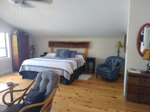 Cozy bedroom with a wooden bed, striped bedding, armchair, and wooden floors, featuring natural light and simple decor.