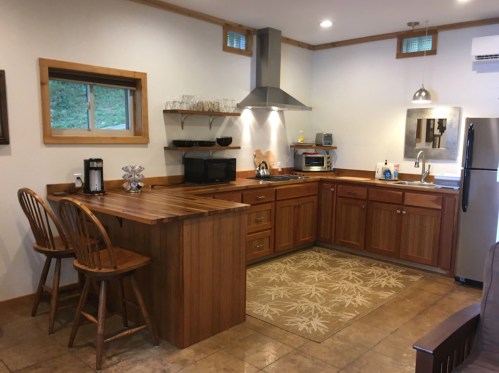 Modern kitchen with wooden cabinets, stainless steel appliances, and a bar with two stools. Natural light from a window.