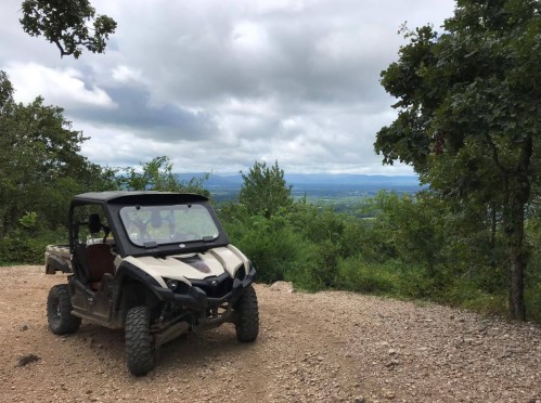 A parked off-road vehicle on a gravel path, surrounded by greenery and a cloudy sky in the background.