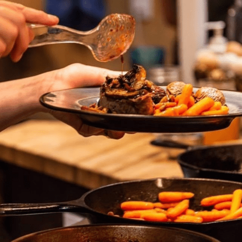 A hand holds a plate of meat with carrots and potatoes, while drizzling sauce over the dish.