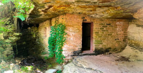 Abandoned stone structure surrounded by greenery, with a partially open door and rocky terrain.