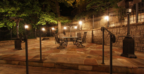 A dimly lit outdoor patio with stone flooring, a table, chairs, and heaters surrounded by trees and a stone wall.