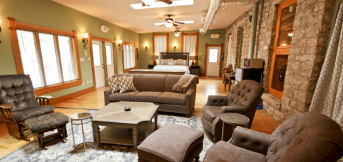 Cozy living area with a sofa, armchairs, and a bed in a well-lit room featuring stone walls and wooden accents.