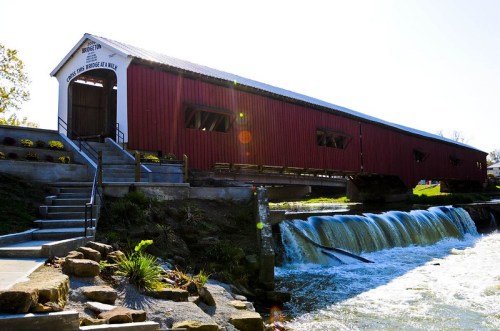 A red covered bridge spans a small waterfall, surrounded by greenery and steps leading down to the water.