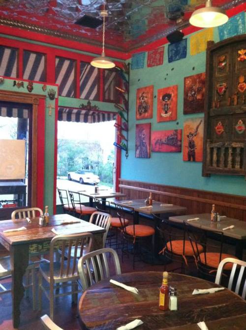 Colorful restaurant interior with wooden tables, chairs, and vibrant wall art. A car is visible outside through the window.